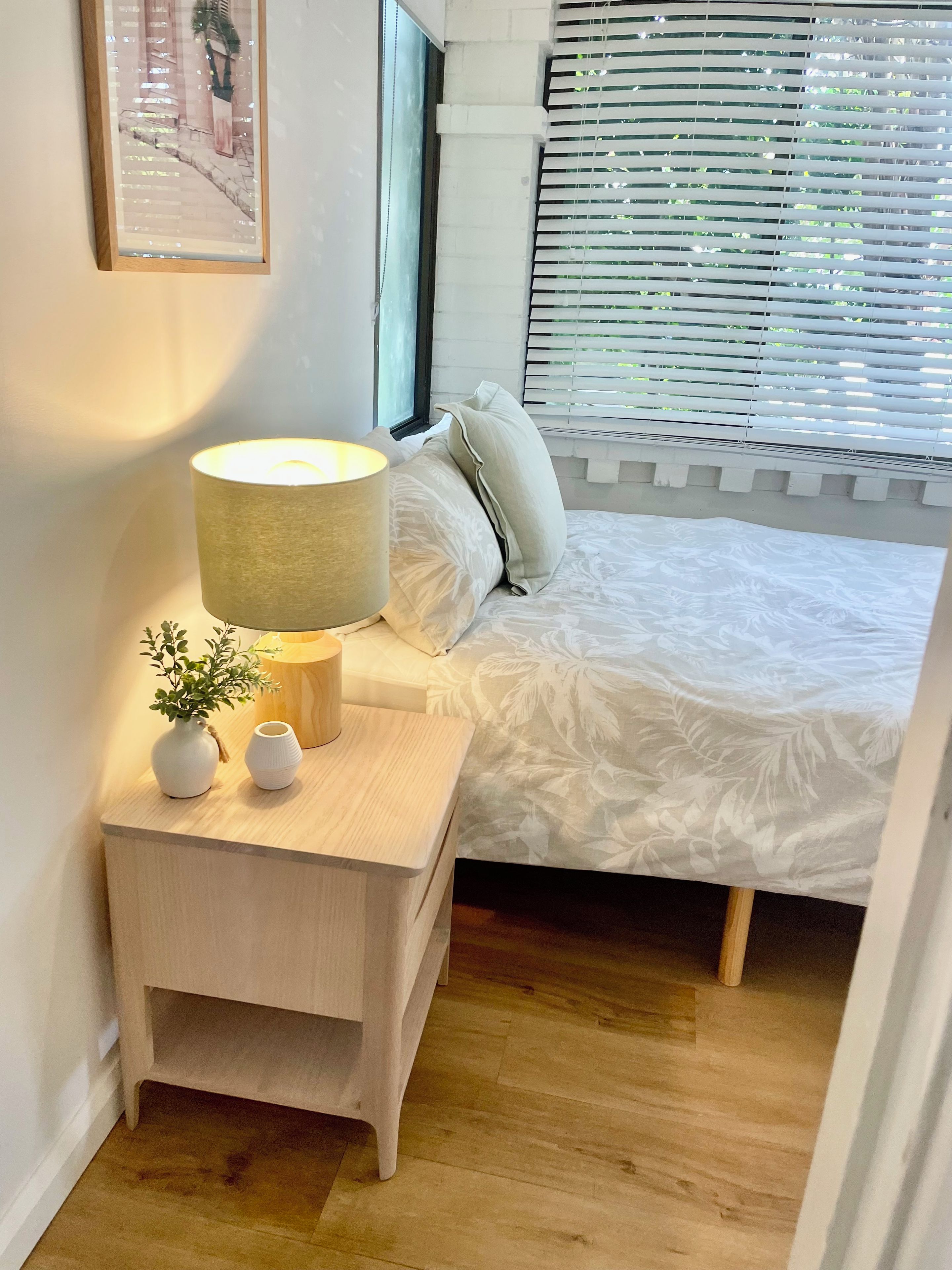 Quarto com cama, mesa de cabeceira com abajur, uma pequena planta e janela com persianas.