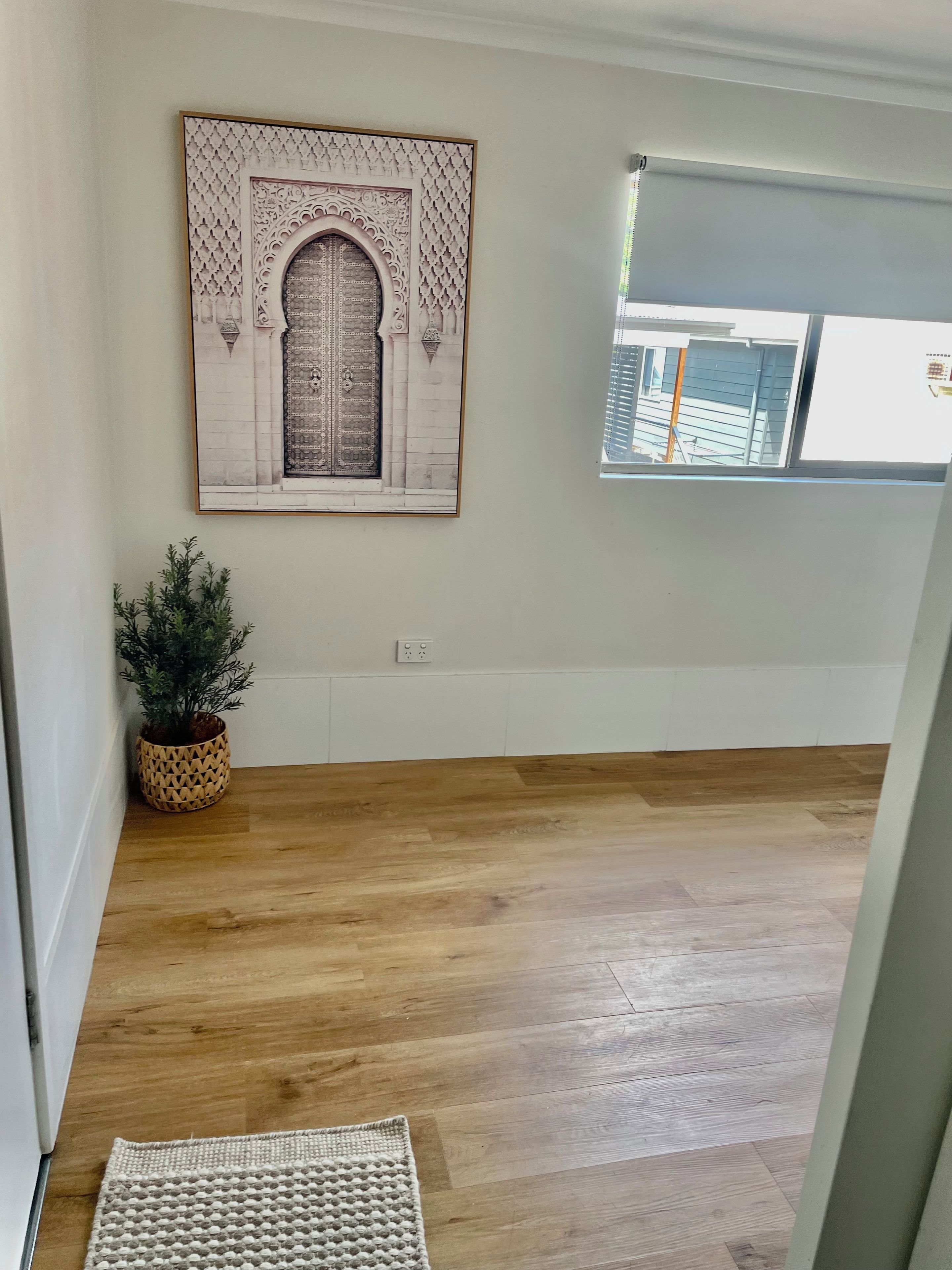 A room with a wooden floor, a framed artwork depicting an ornate door, a potted plant, and a window with a view of buildings.