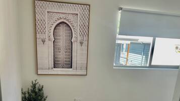 A room with a wooden floor, a framed artwork depicting an ornate door, a potted plant, and a window with a view of buildings.