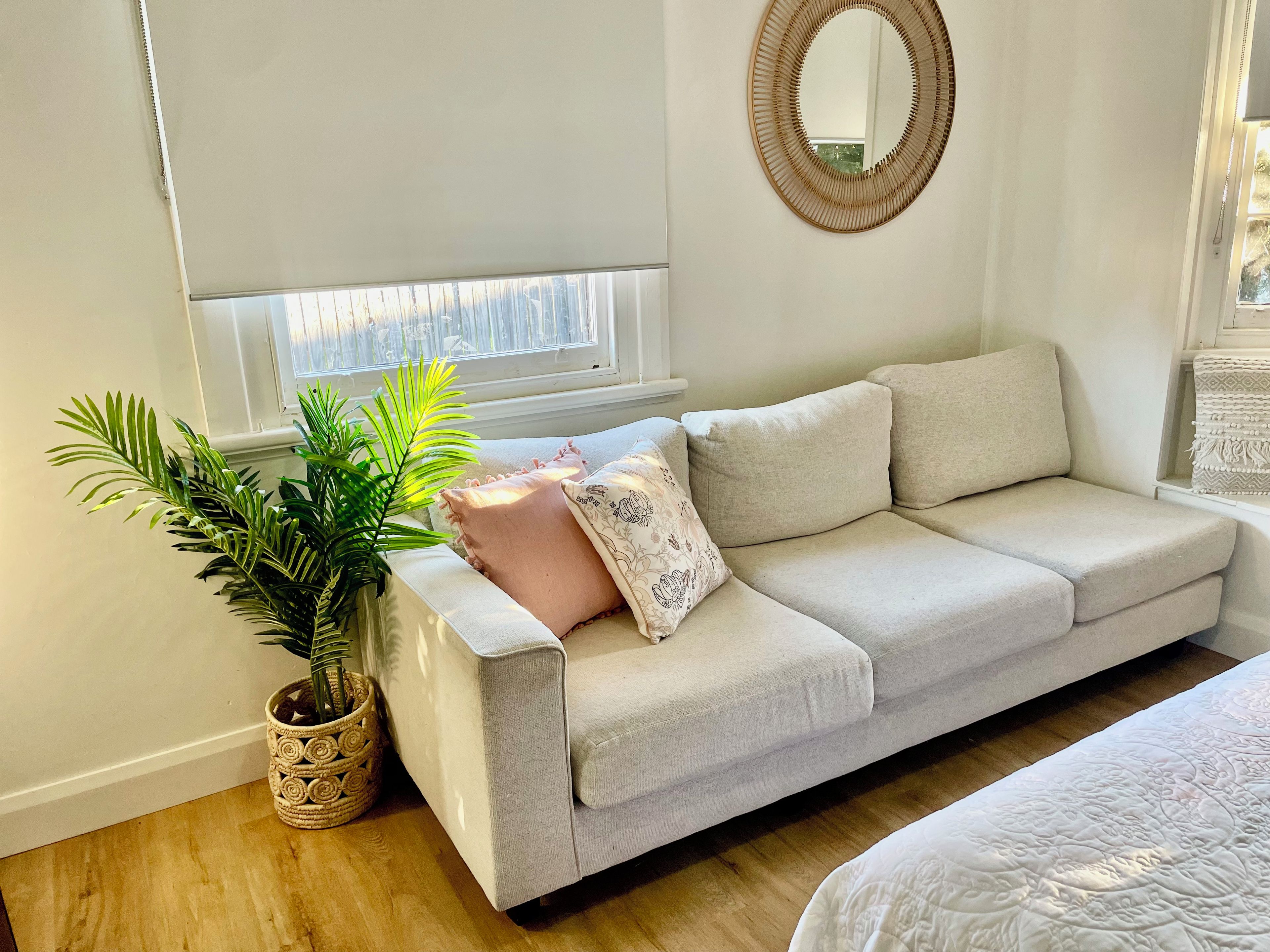 A room with a light-colored sofa, a potted plant, a mirror, and a window with blinds.