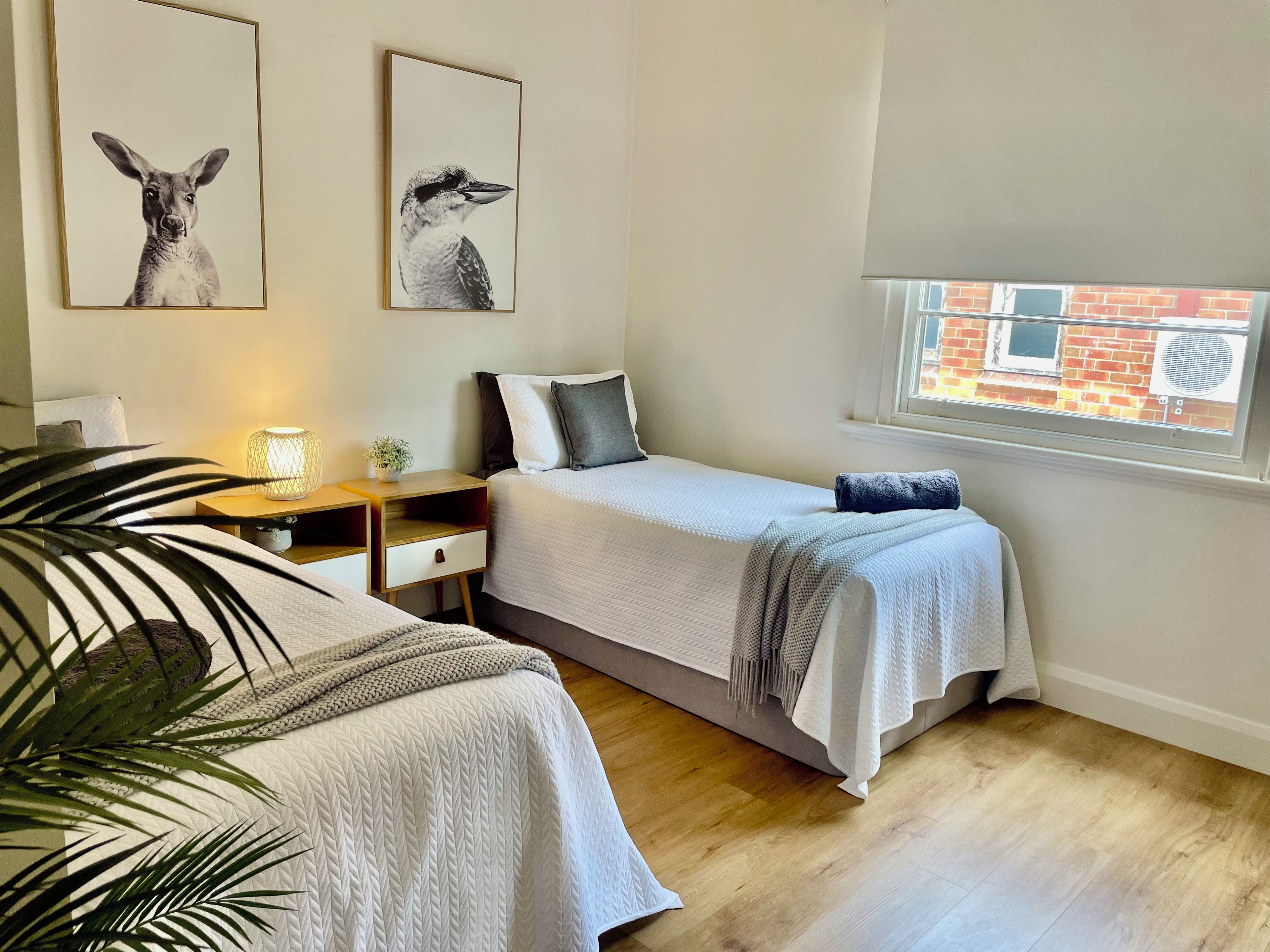 A bedroom with two beds, a bedside table with a lamp, a window with a view of a brick building, and two wall-mounted animal prints.