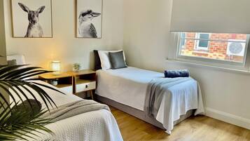 A bedroom with two beds, a bedside table with a lamp, a window with a view of a brick building, and two wall-mounted animal prints.
