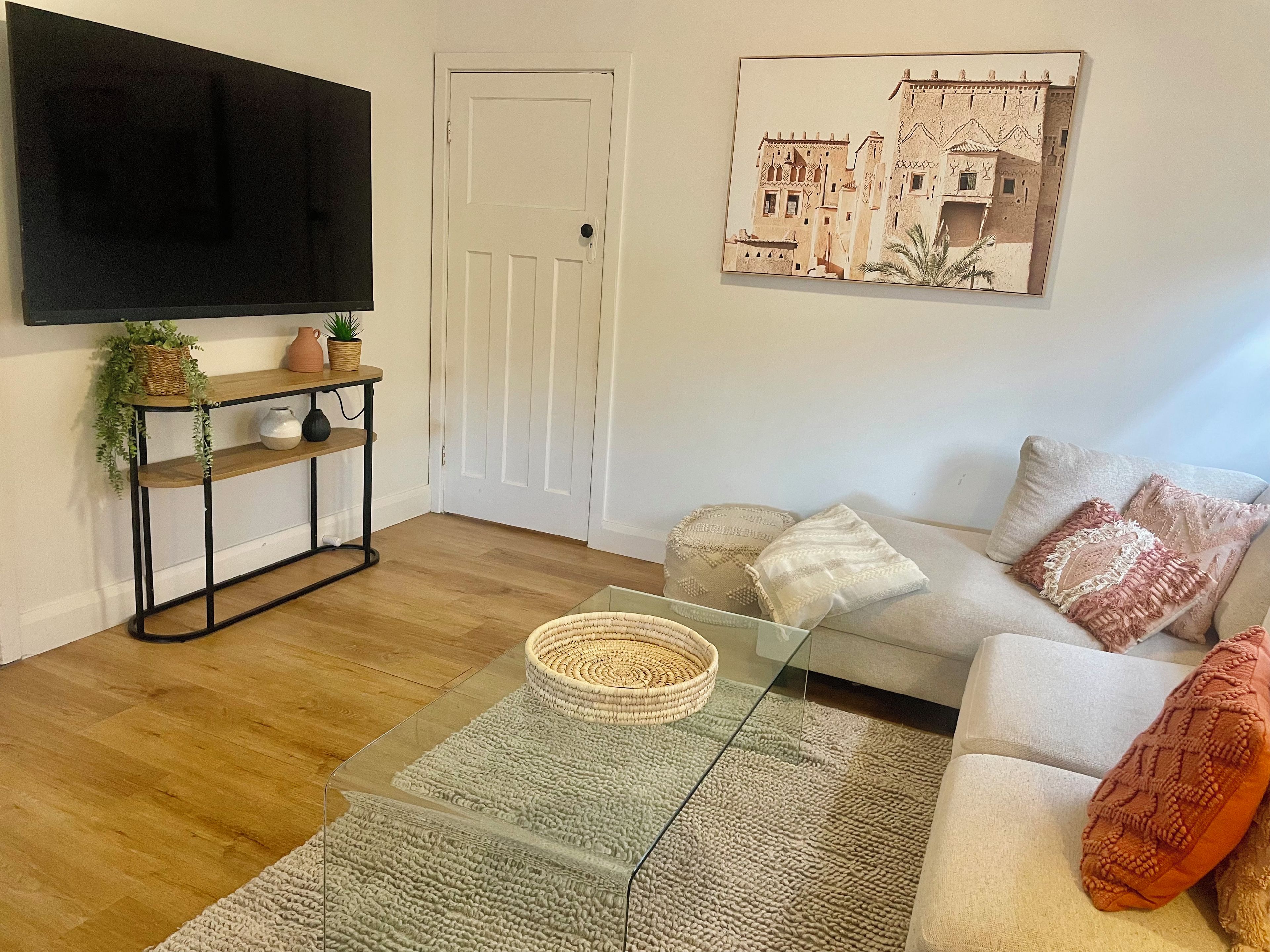 A living room with a flat-screen TV, a sofa with patterned cushions, a glass coffee table, and a wall-mounted picture.