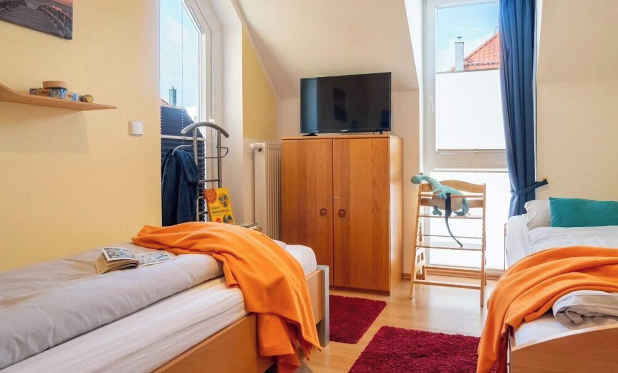 A bedroom with two beds, a wardrobe, a television, and a window with a view of buildings.