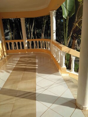 A covered outdoor area with a tiled floor, white balustrades, and a view of greenery.
