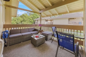 Un espace extérieur aménagé, abrité et doté d’un canapé, d’un pouf et de fauteuils, ainsi que d’une petite table avec un vase, offrant une vue sur une maison en arrière-plan.