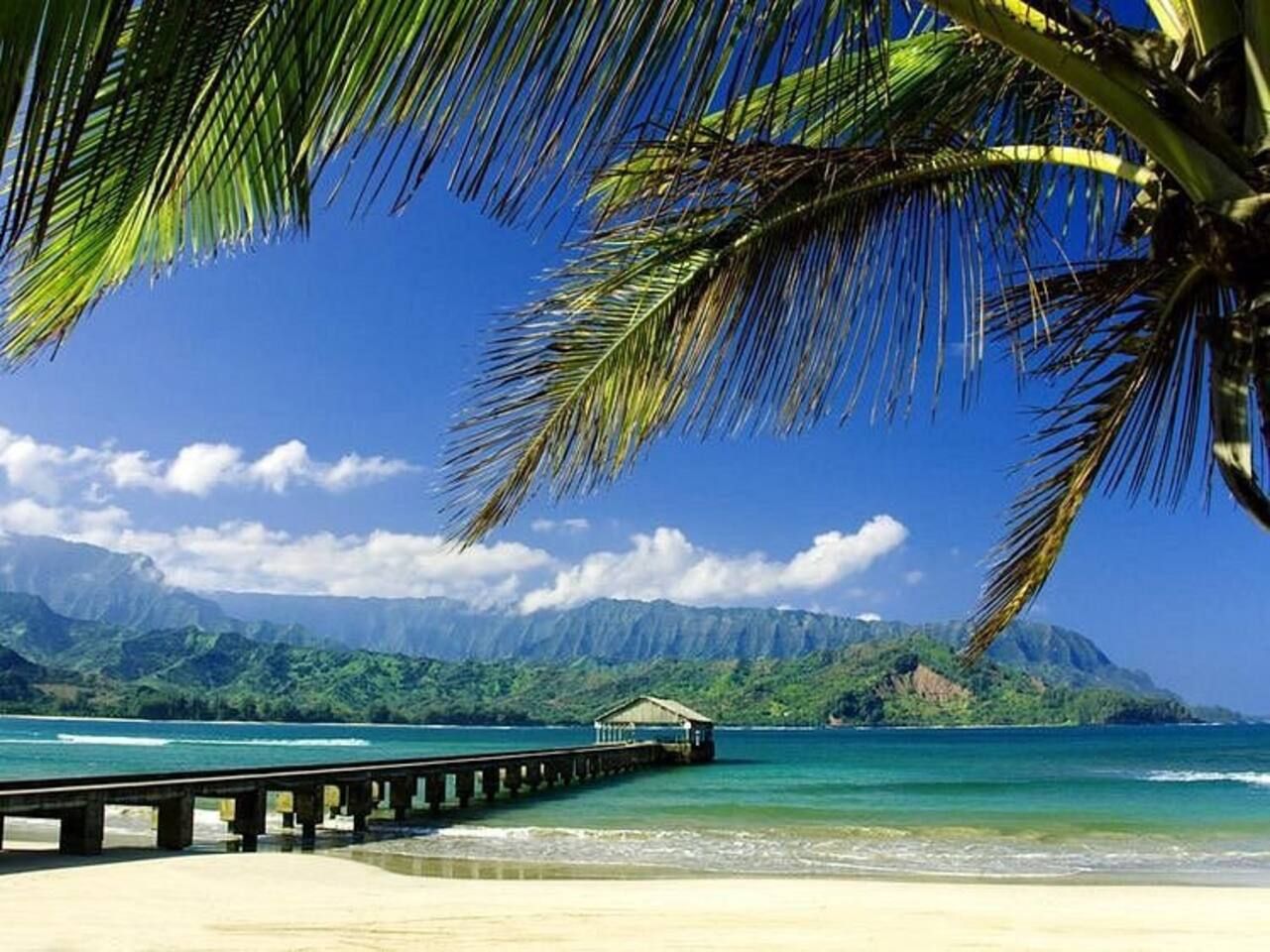 Ein Strand mit einem hölzernen Steg, der zu einer kleinen Hütte führt, umgeben von klarem, blauem Wasser und grünen Bergen im Hintergrund.