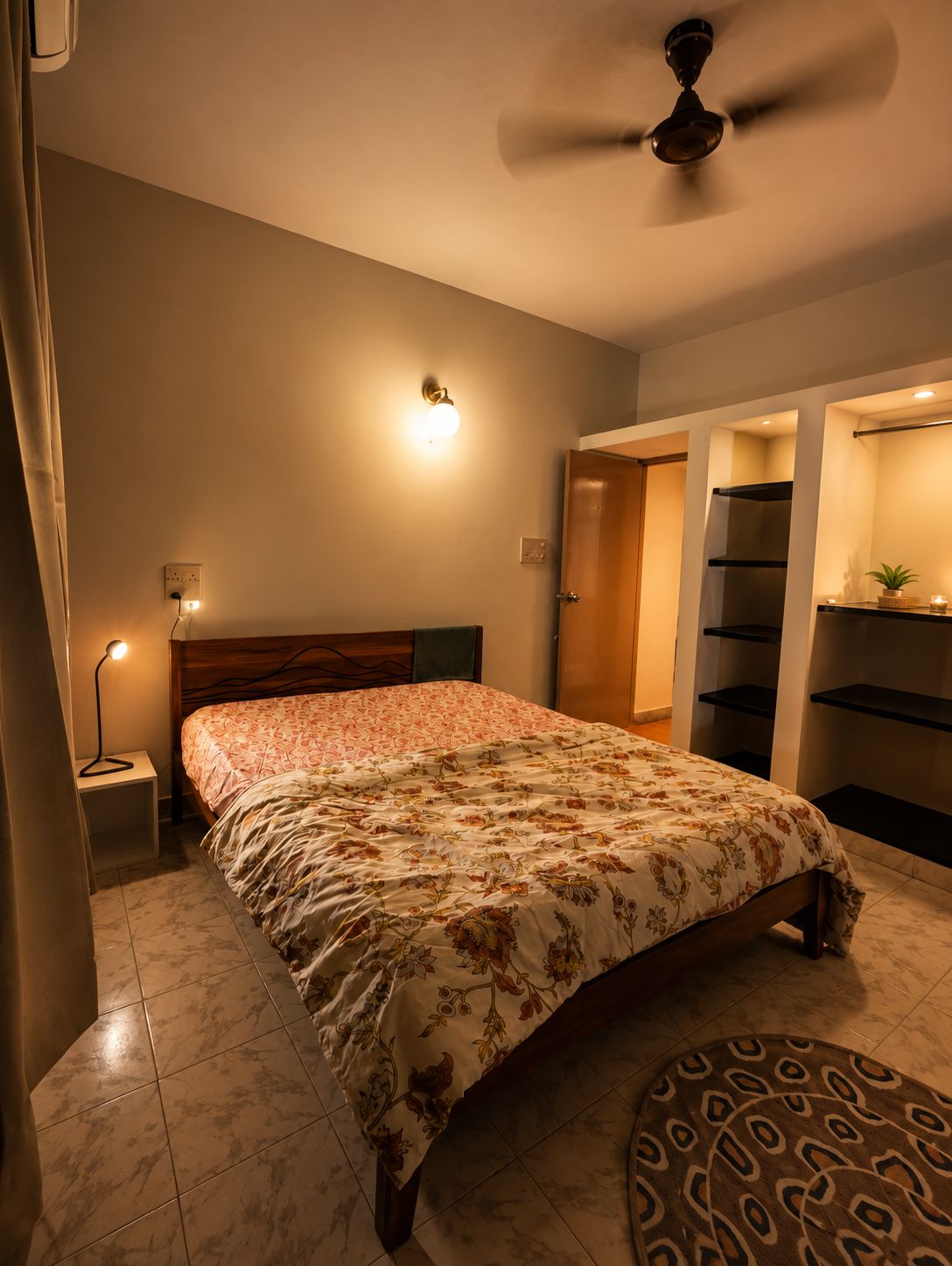 A bedroom with a bed, a ceiling fan, a nightstand with a lamp, and a wall-mounted shelf.