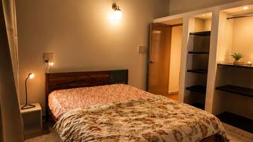 A bedroom with a bed, a ceiling fan, a nightstand with a lamp, and a wall-mounted shelf.