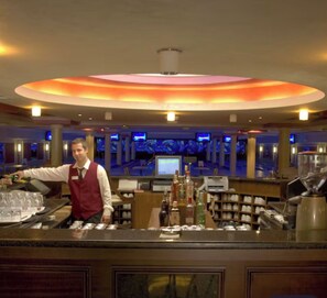 Zona de bar con un bartender, una barra con vasos y botellas, y un fondo con varias pantallas.