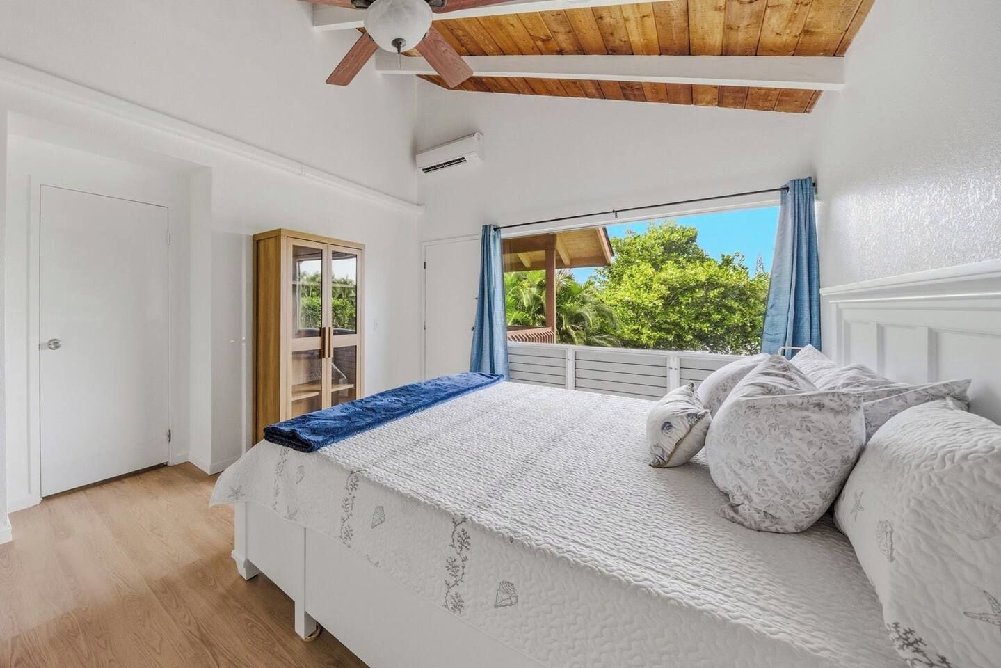 Ein Schlafzimmer mit einem Bett, einem Deckenventilator, einem Fenster mit Vorhängen und Blick ins Grüne.
