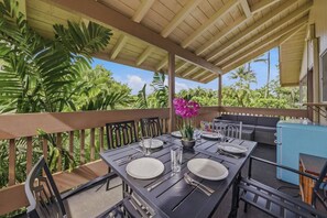 Un área de comedor al aire libre con una mesa dispuesta para dos personas, rodeada de sillas, un enfriador azul y una planta en maceta con flores rosadas.