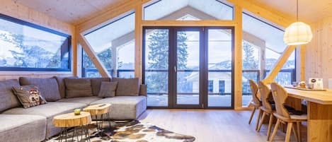 A cozy cabin interior with a wooden ceiling, large windows, a sofa, a dining table, and a cowhide rug.