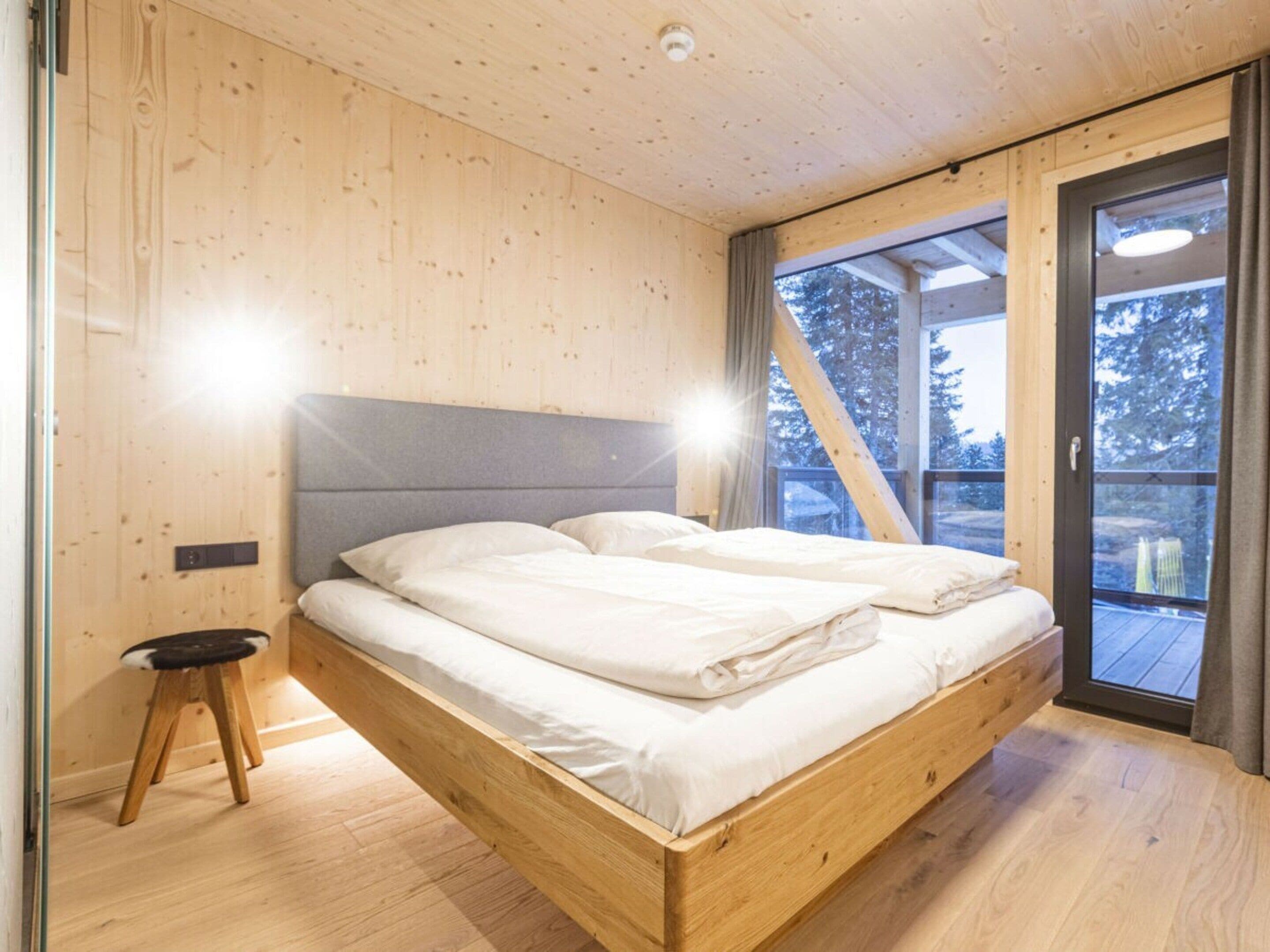 Une chambre à coucher en bois, avec un lit, un petit tabouret et une fenêtre donnant sur des arbres.