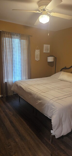 A bedroom with a large bed, a ceiling fan, a bedside lamp, and a window with curtains.