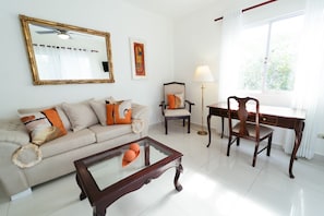 A living room with a sofa, a coffee table, a desk with a chair, a floor lamp, a large mirror, and a window with curtains. - Beautiful 3-bedroom apartment,3 Min walk to the Beach,Costambar Puerto Plata,DR (Puerto Plata)