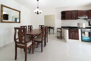 A dining area with a wooden table and chairs, a chandelier, and a kitchen with dark wooden cabinets and a blue appliance. - Beautiful 3-bedroom apartment,3 Min walk to the Beach,Costambar Puerto Plata,DR (Puerto Plata)