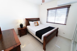 A bedroom with a wooden bed, a bedside table with a lamp, a window with blinds, and a tiled floor. - Beautiful 3-bedroom apartment,3 Min walk to the Beach,Costambar Puerto Plata,DR (Puerto Plata)