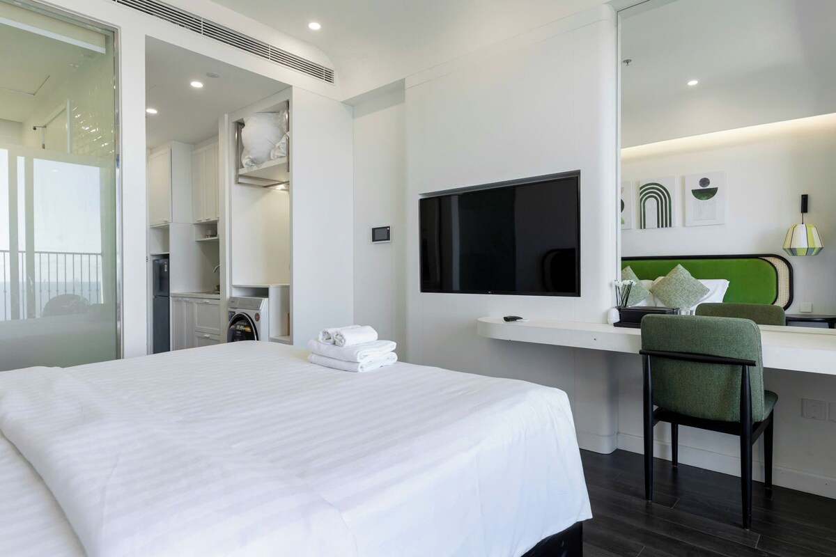 A hotel room with a bed, a desk with a flat-screen TV, a chair, and a green headboard.