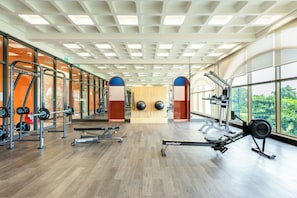 A gym with cardio equipment, weightlifting stations, and large windows offering views of greenery. - The Clound Apartment Hillside Phu Quoc (Phu Quoc)