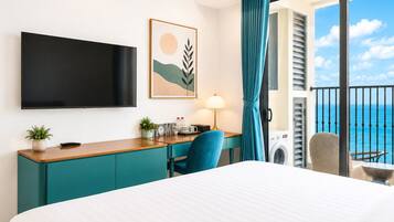A hotel room with a bed, a desk with a lamp, a TV, a view of the ocean, and a balcony with a chair.