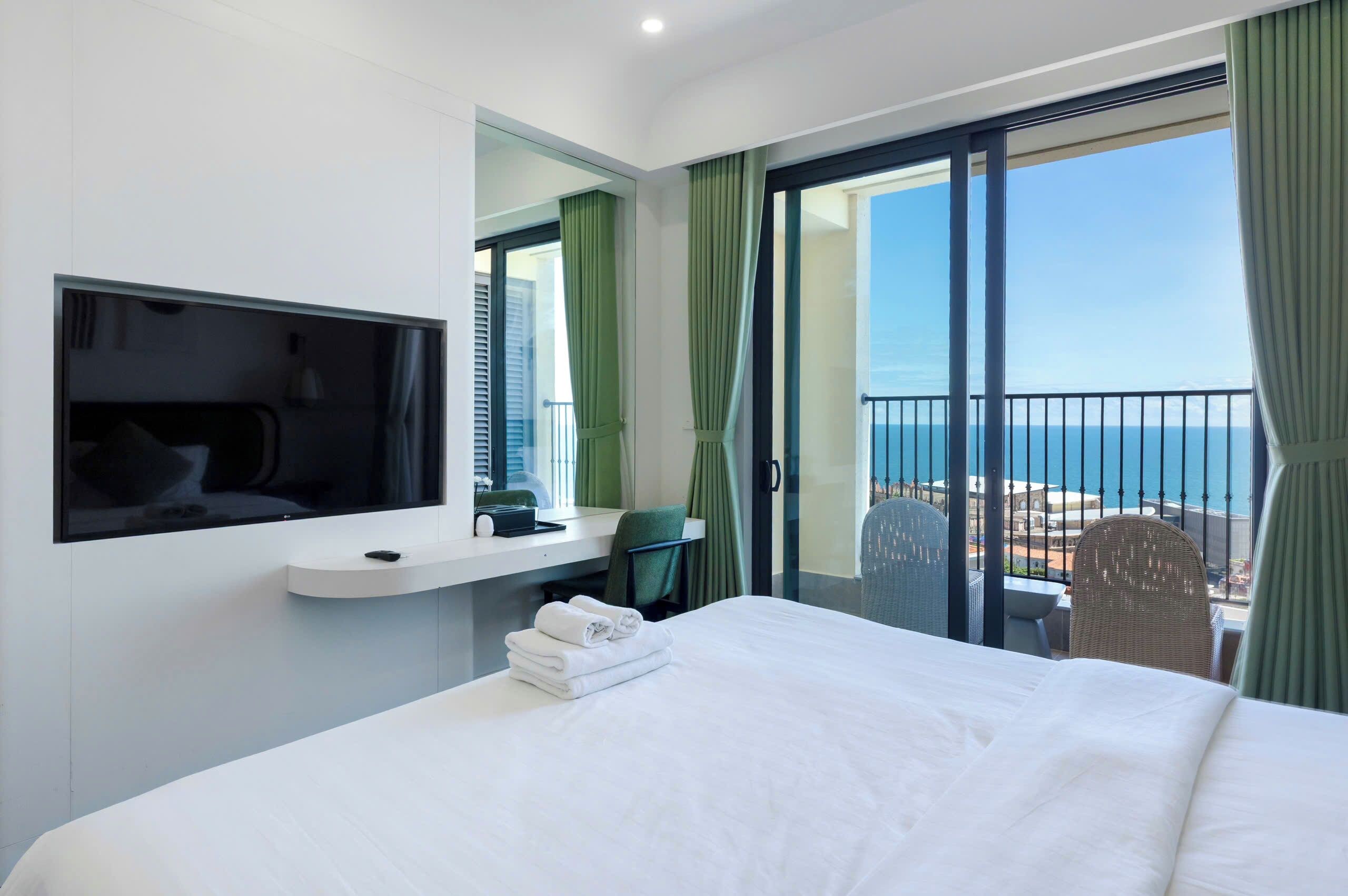 A hotel room with a bed, a TV, a desk, a chair, and a balcony with a view of the sea.