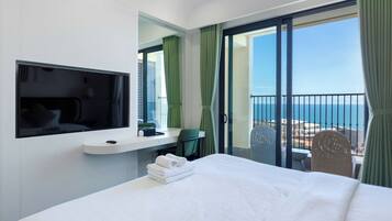 A hotel room with a bed, a TV, a desk, a chair, and a balcony with a view of the sea.