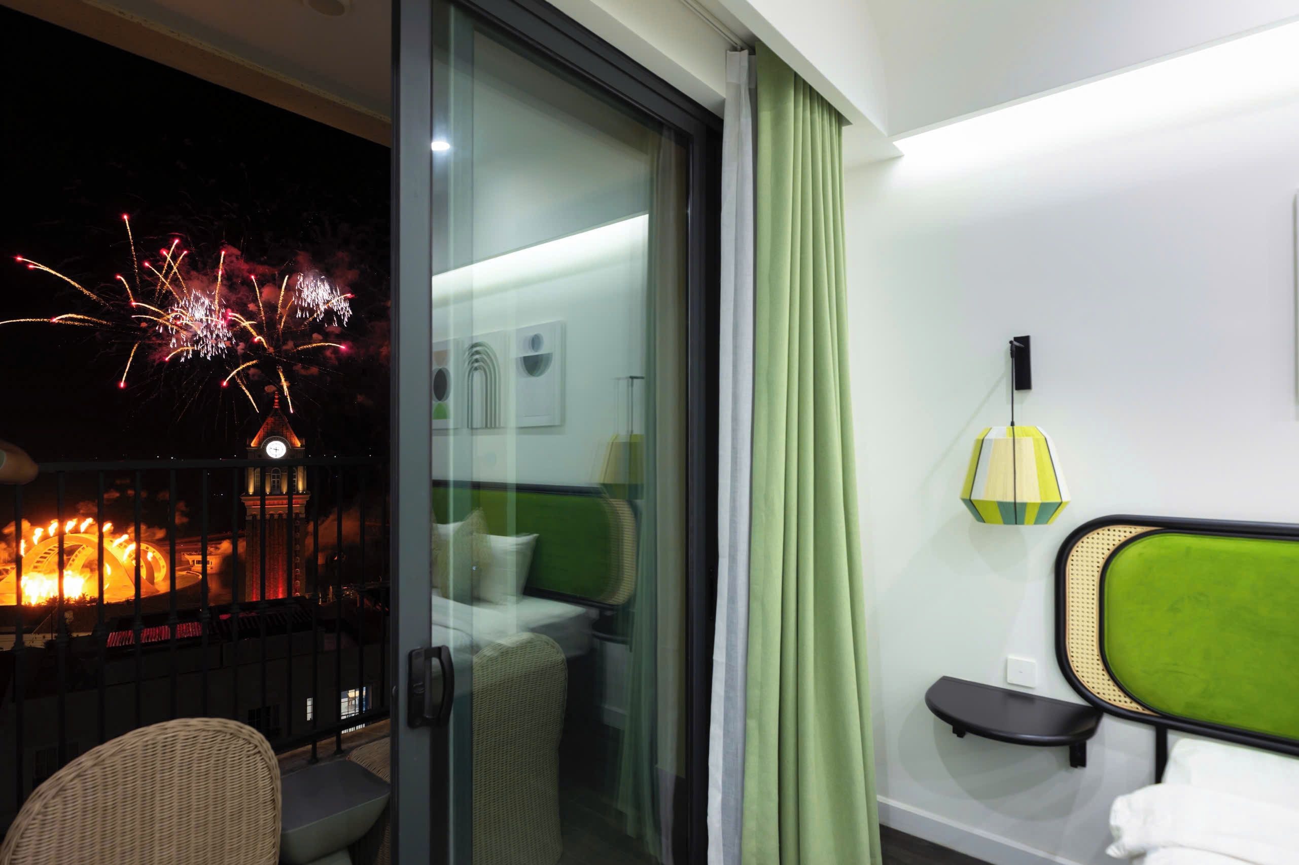 Una camera d'albergo con balcone, un letto con testiera verde, un tavolino e una lampada a parete.