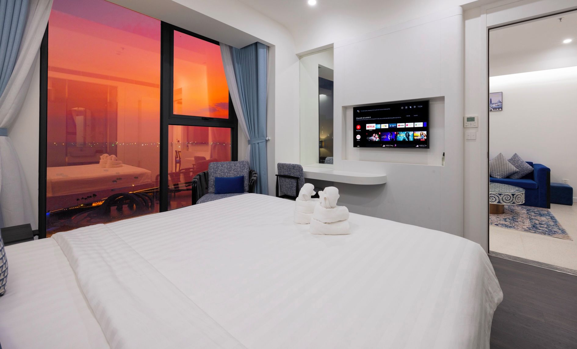 Camera d'albergo con un letto grande, una TV montata a parete, un'area salotto con divano e poltrone, e vista sul tramonto dalla finestra.