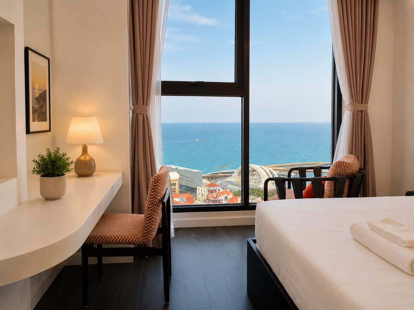 A hotel room with a desk, chair, bed, and a view of the sea and cityscape.