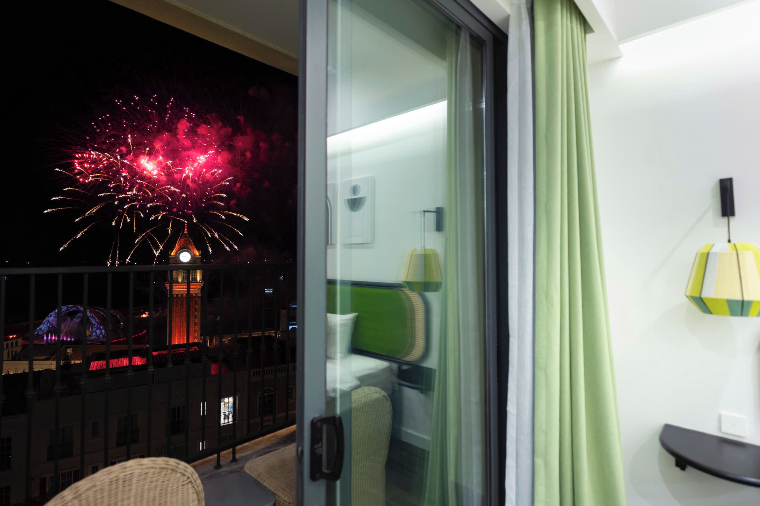 A balcony with a view of fireworks, a glass door, a bed, a lamp, and a chair.