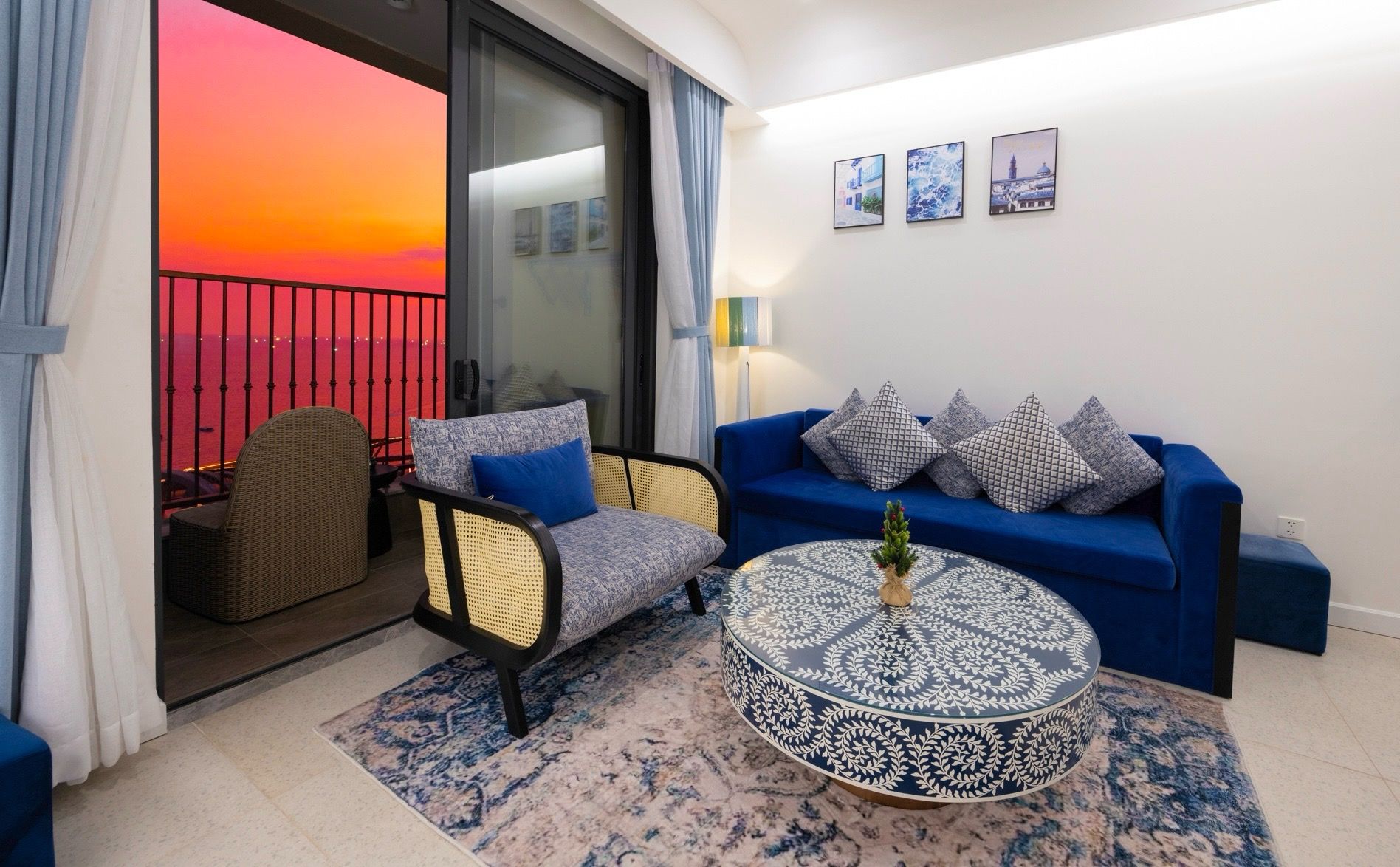 A living room with a view of a sunset over the sea, a blue sofa, a wicker chair, and a patterned coffee table.
