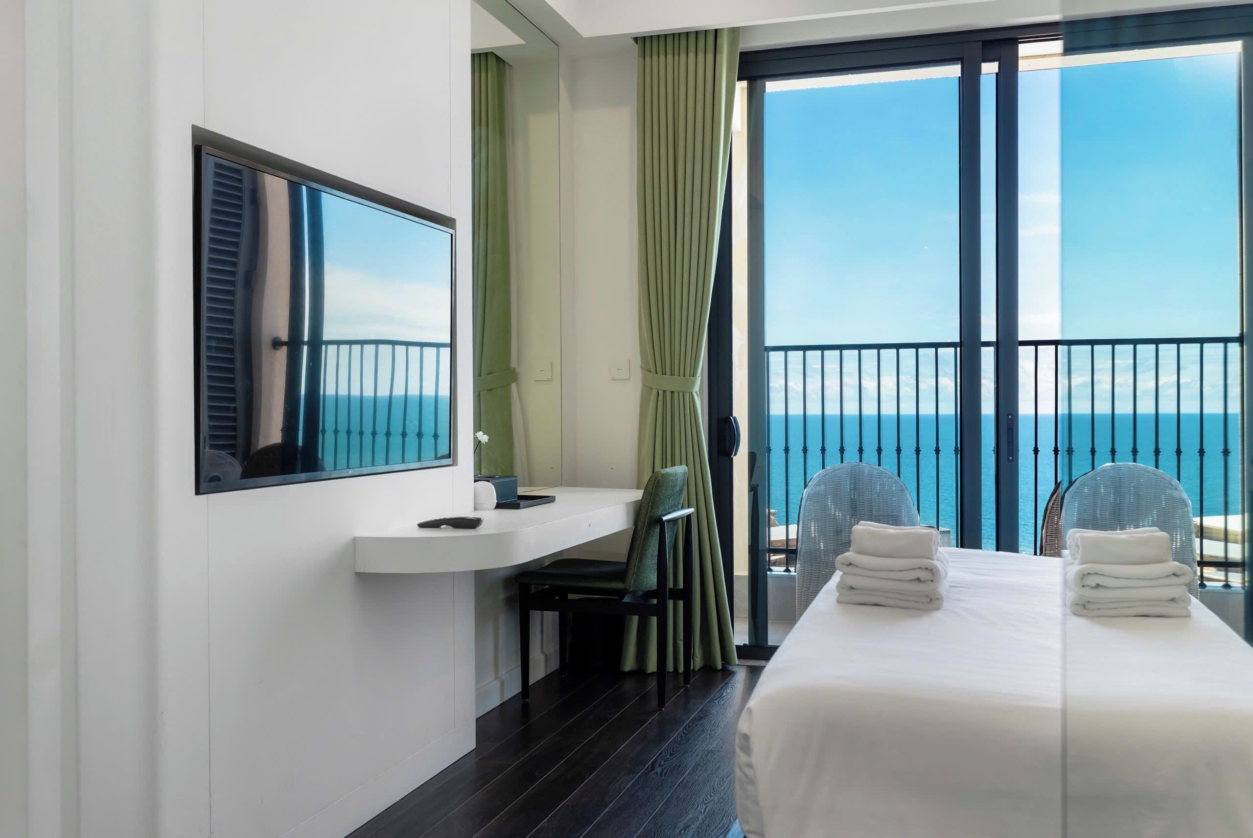 A hotel room with a bed, a desk, a chair, a window with a view of the sea, and a balcony with a railing.