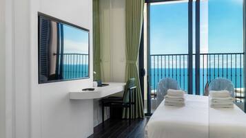 A hotel room with a bed, a desk, a chair, a window with a view of the sea, and a balcony with a railing.