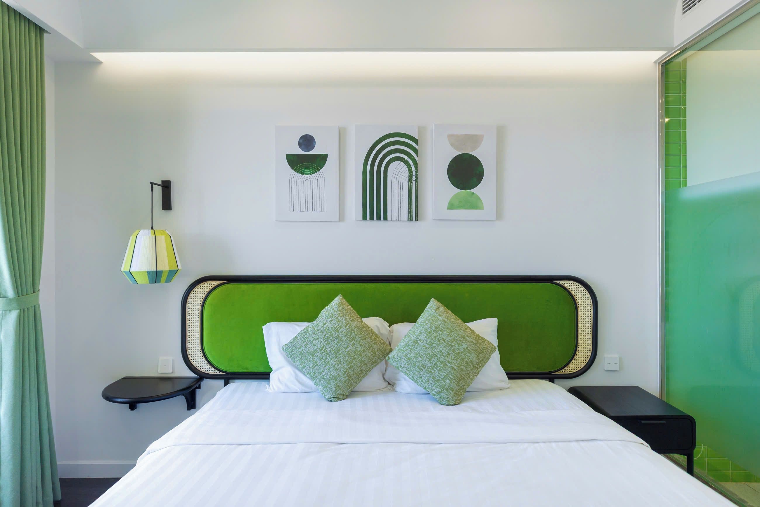 A modern hotel room with a green headboard, a bed with white linens, two green pillows, a bedside table with a lamp, and three abstract wall art pieces.