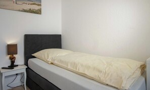 Ein Einzelbett mit grauem Kopfteil und weißer Matratze, ein Nachttisch mit einer Lampe sowie ein Bild an der Wand.