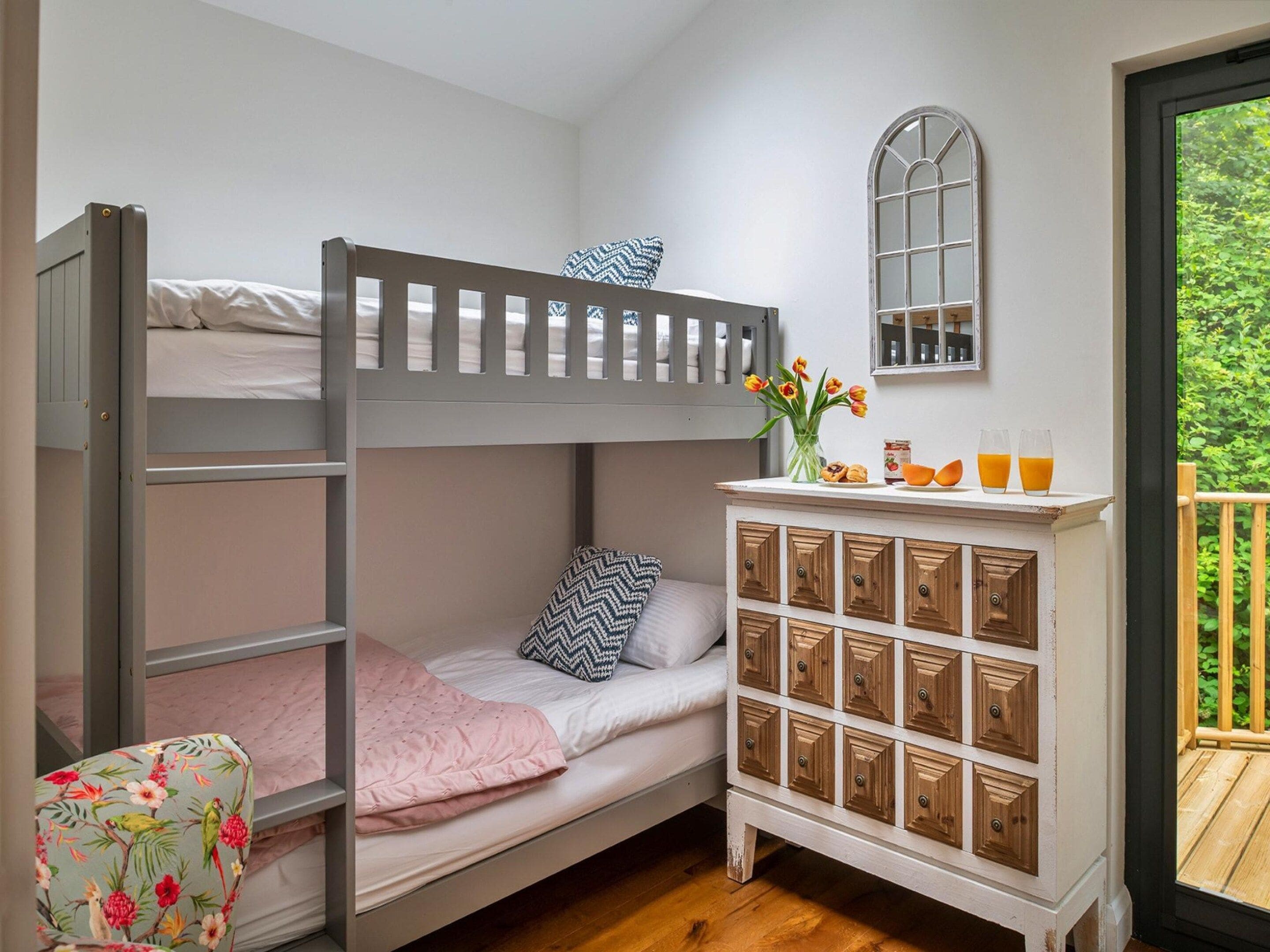 A bunk bed with a white headboard, a small bedside table with a vase of flowers, and a window with a view of greenery.