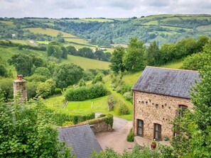 A stone building with a sloped roof, surrounded by lush greenery and rolling hills in the background. - Luxury Barn With Private Hot Tub (Ilfracombe)
