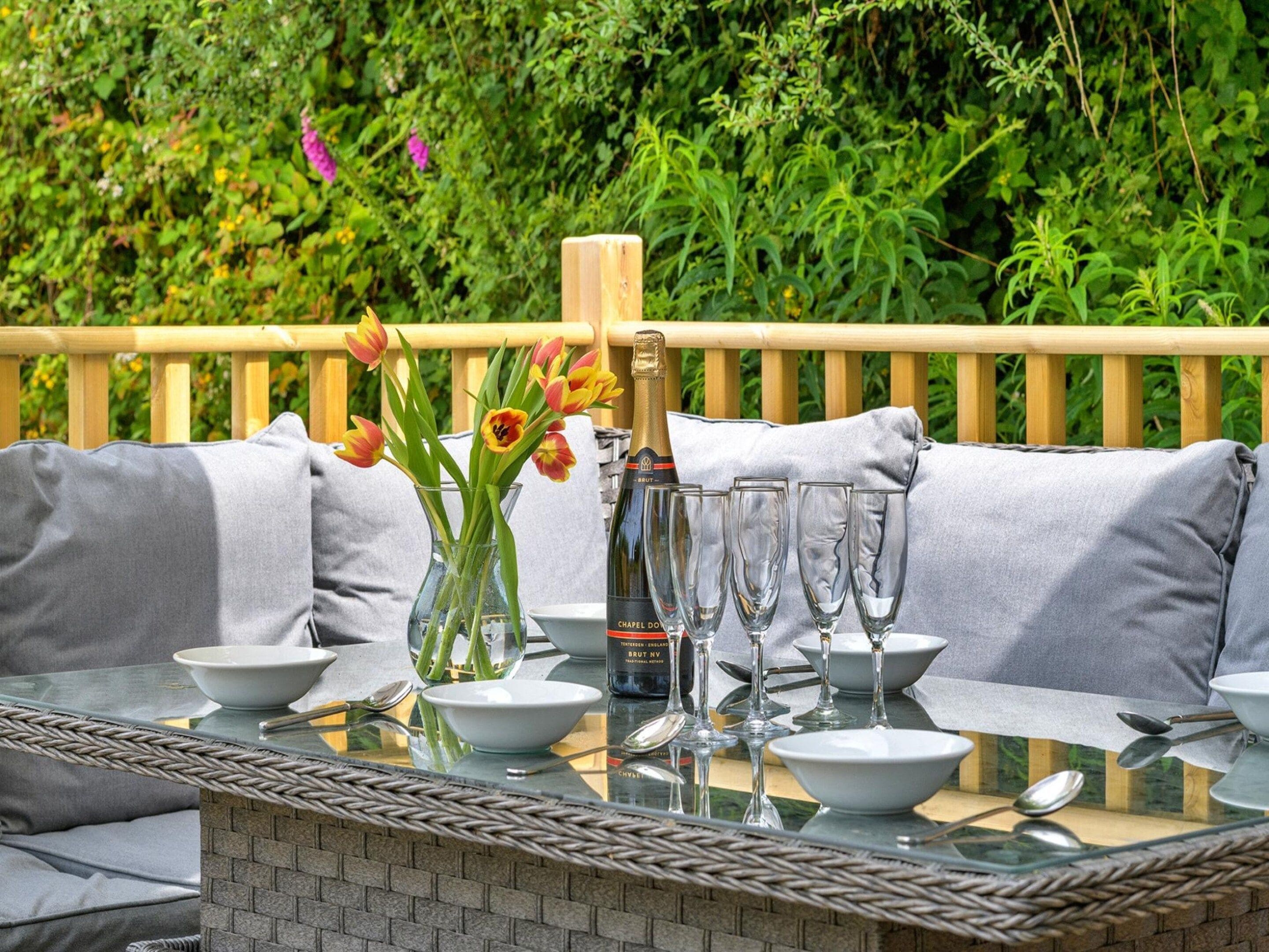 An outdoor dining area with a glass table, grey cushioned seating, and a bottle of champagne.