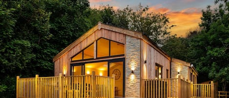 A wooden cabin with a hot tub, surrounded by a wooden fence, set against a backdrop of trees and a sunset sky.