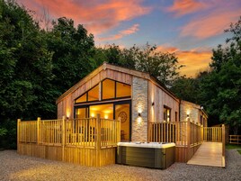 Una cabaña de madera con jacuzzi, rodeada por una cerca de madera, con árboles y un cielo de atardecer de fondo.