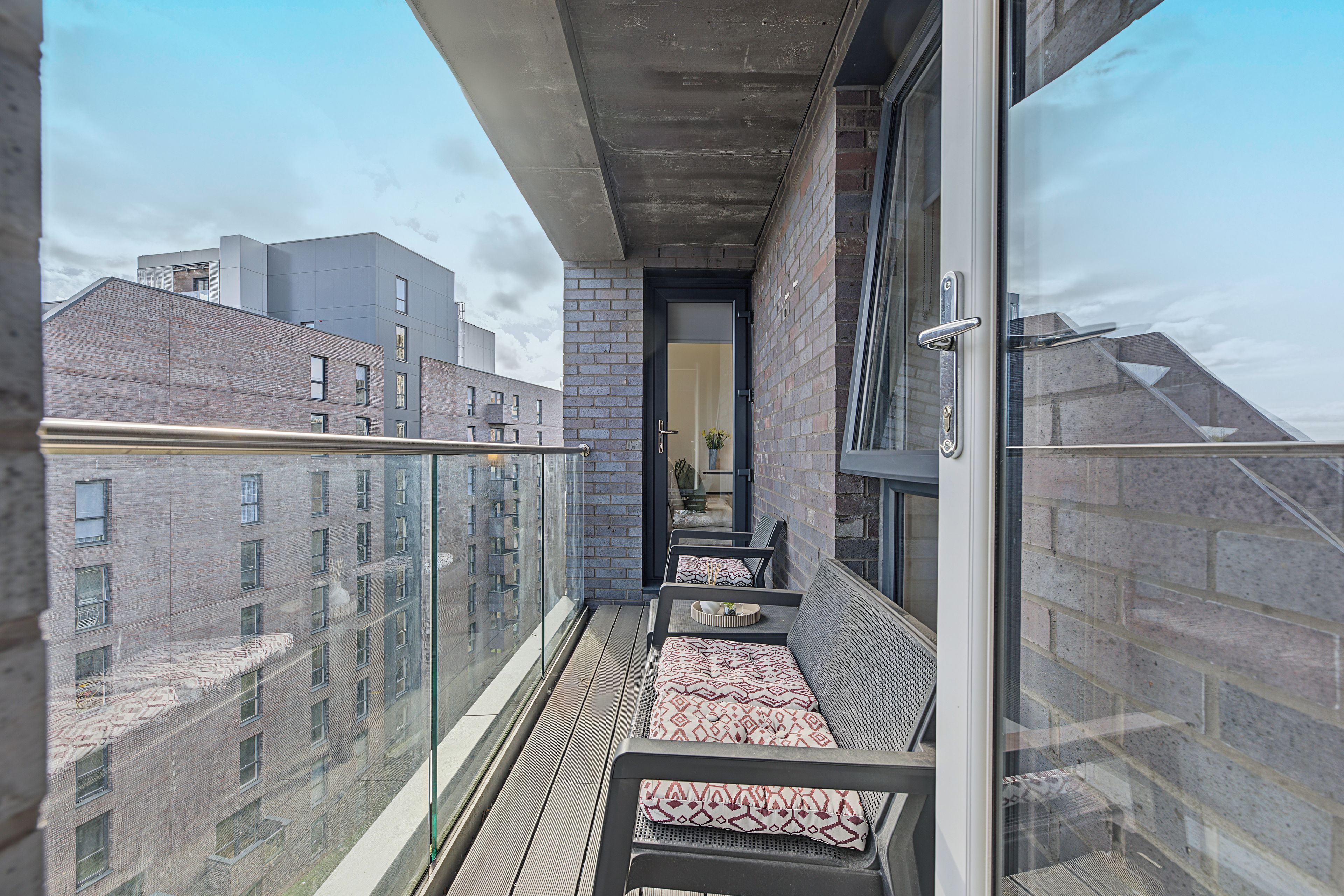 A balcony with a glass railing, a seating area with patterned cushions, and a view of adjacent buildings.