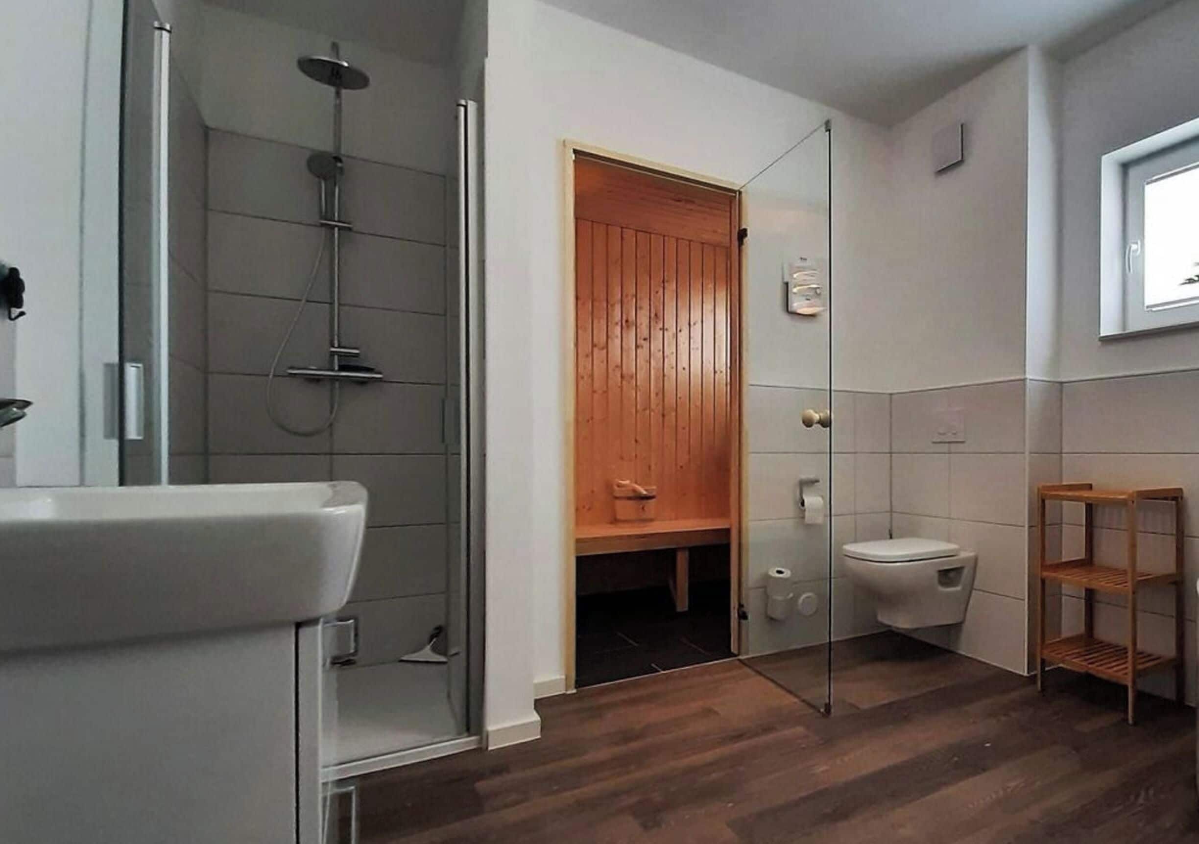 A bathroom with a glass shower enclosure, a white sink, a wooden door, a toilet, and a shelf.