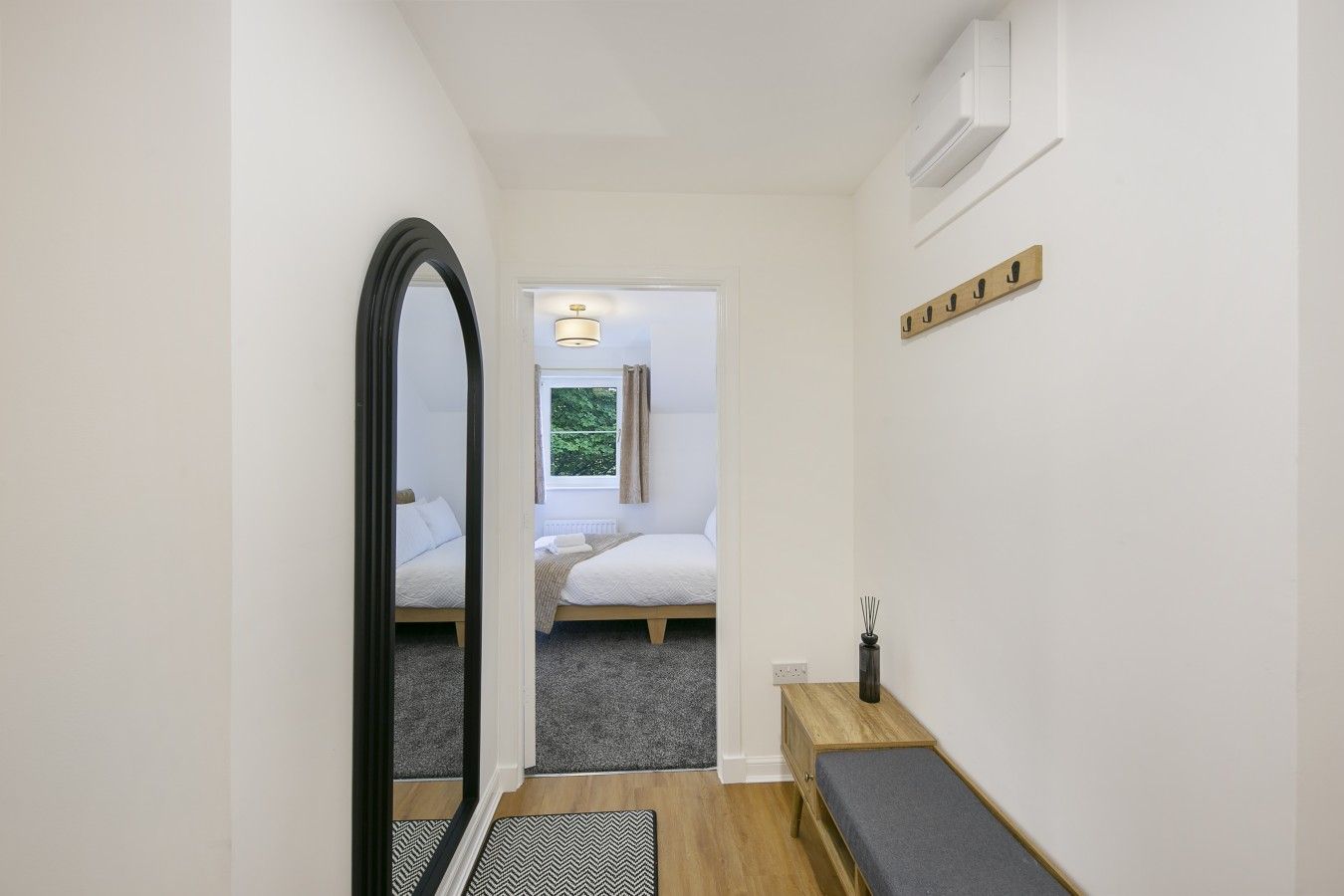 Een smalle gang die uitkomt in een slaapkamer met een bed, een bankje en een spiegel.