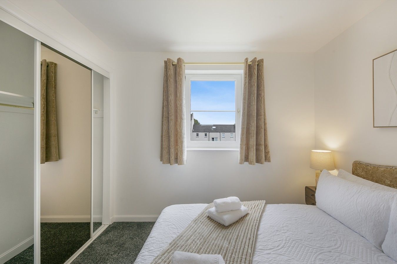 Una habitación de hotel con cama, ventana con cortinas, lámpara de noche y vistas a edificios.
