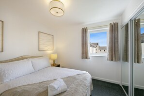 A hotel room with a bed, a bedside lamp, a window with curtains, and a view of buildings outside. - Eday Lodge - Grampian Lettings (Aberdeen)