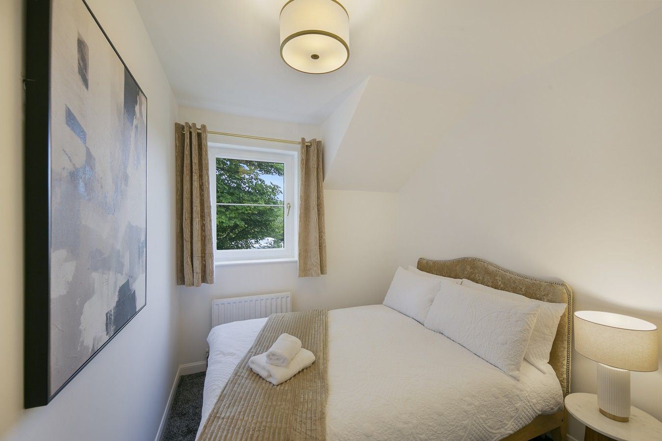 Een slaapkamer met een bed, een raam met gordijnen, een radiator, een wandlamp en een ingelijst schilderij.