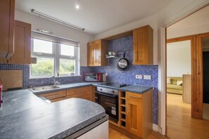 A kitchen with wooden cabinets, a built-in oven, a microwave, and a window with a view of greenery. - Lea Cottage - Spacious 2 bed cottage near Lochgilphead (Cairnbaan)