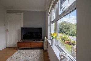 A living room with a television, a radiator, a window with a view of greenery, and a door. - Lea Cottage - Spacious 2 bed cottage near Lochgilphead (Cairnbaan)