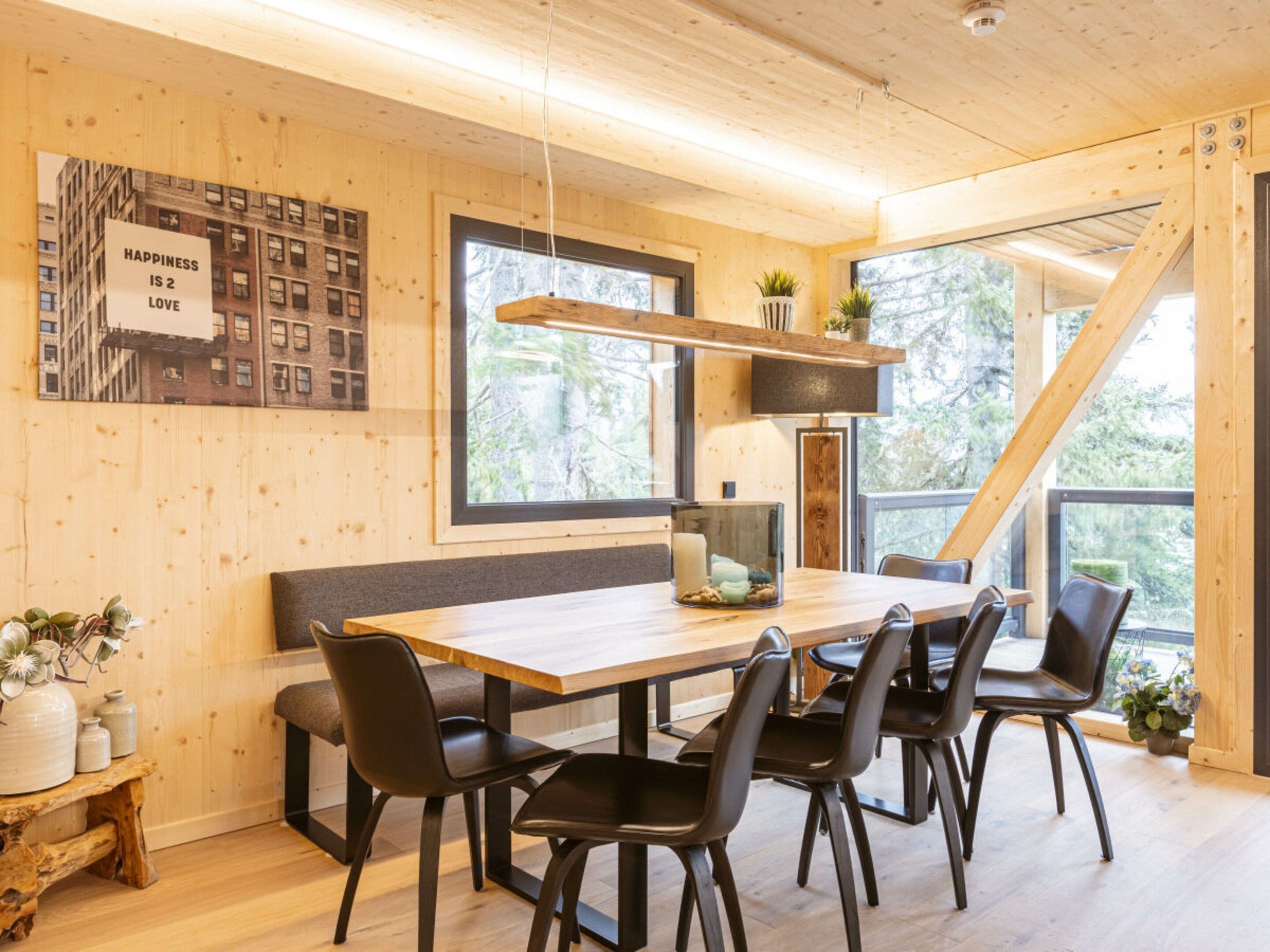 A modern dining area with a wooden table, black chairs, and a large window offering a view of trees.
